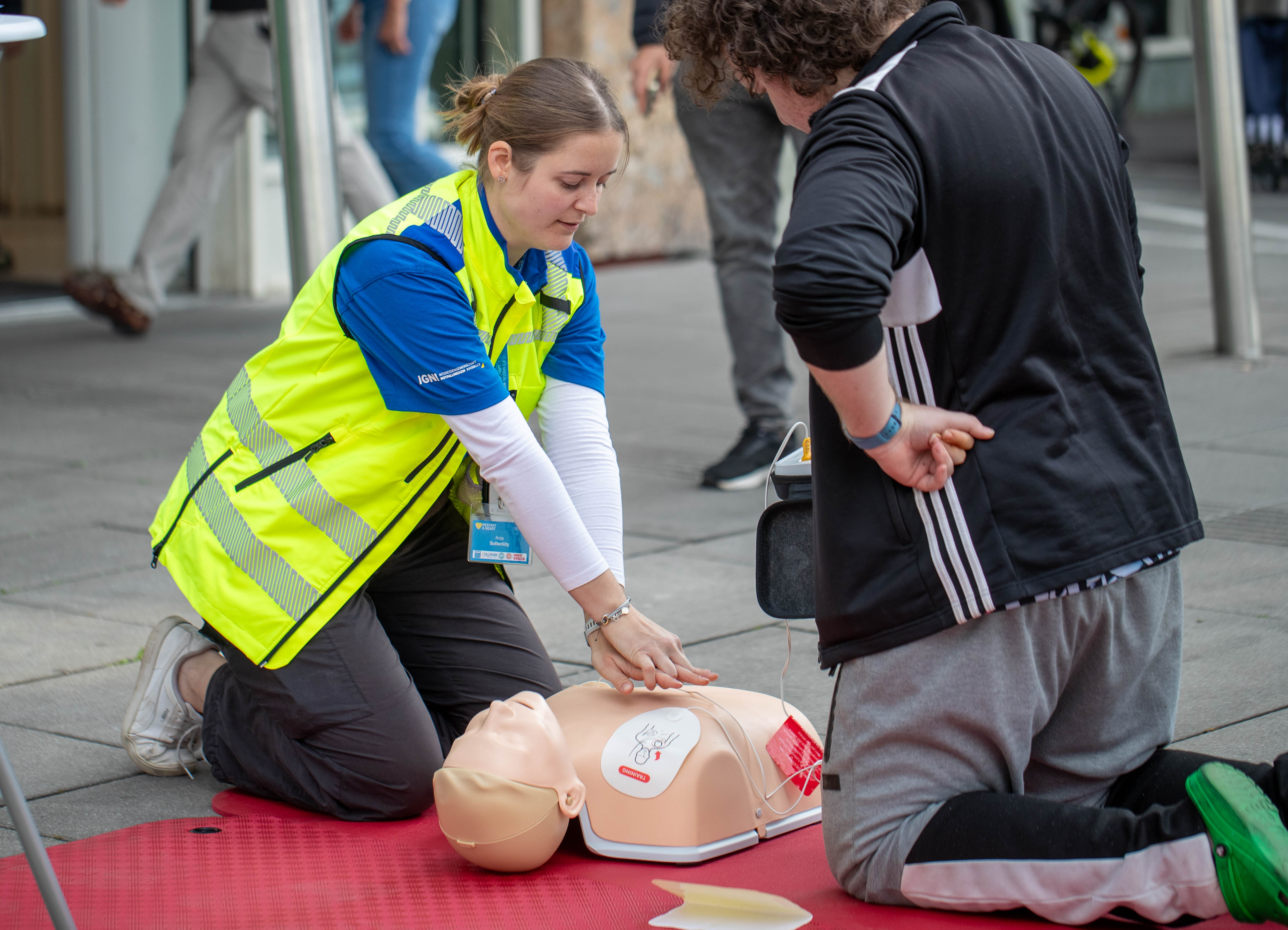 Eine IGNI-Trainerin zeigt einem Teilnehmer Herzdruckmassage und AED-Anwendung an einer Übungspuppe